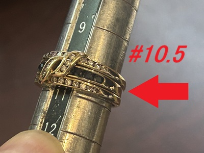 金属製リングゲージにはめられたゴールドの3連インターロッキング指輪。中央のバンドにはサファイア、上下のバンドにはダイヤモンドがセットされている。ゲージは10.5号を指しており、その横に赤い矢印と「#10.5」の赤字テキストがある。